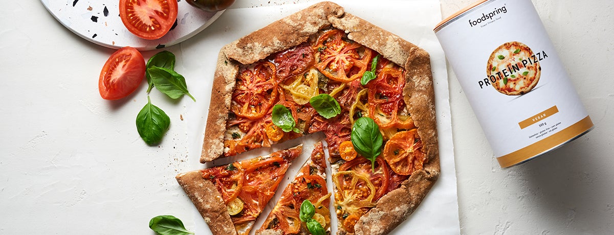 A vegan tomato galette topped with bright red and yellow tomato slices and garnished with fresh basil leaves sits on a white background. In one corner of the image is a canister of foodspring's Protein Pizza mix
