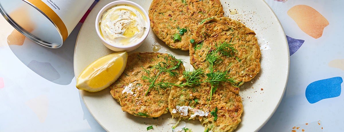 A plate of vegan zucchini pancakes topped with fresh green herbs. A quarter lemon and a bowl of cream-colored dip sit on the side.