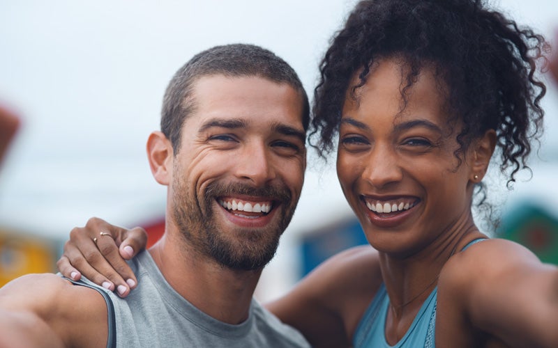 A white man and a woman of color put their arms around each other and smile for a selfie.