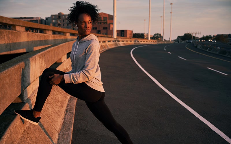 A woman of color stretches on an outdoor highway. The sunset lights her in gold as she finishes her warm up and cool down for runners.