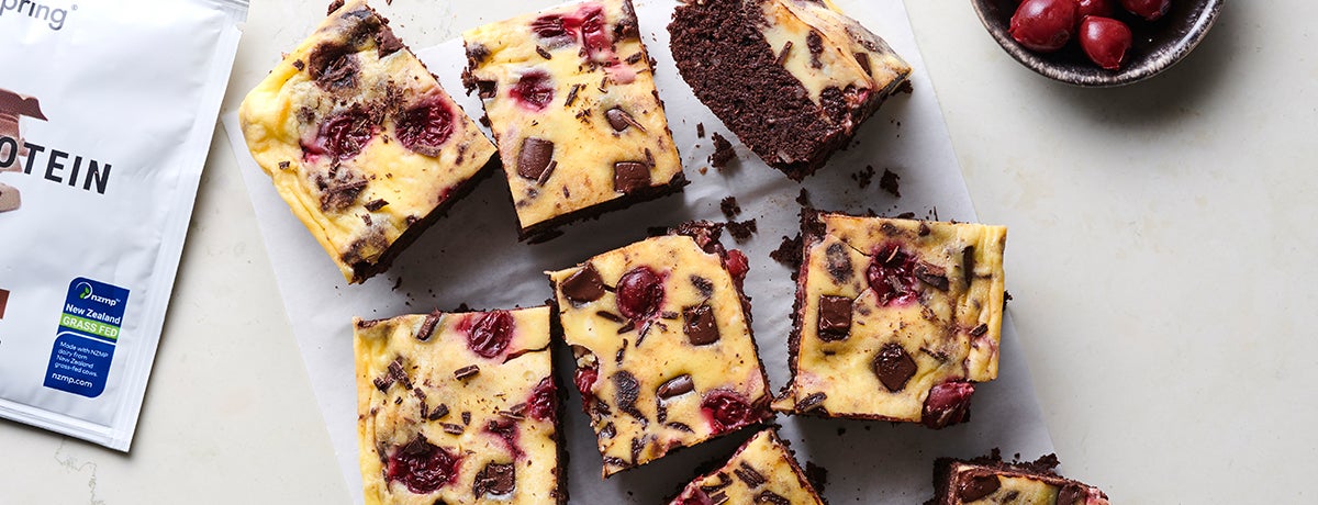 A photo from above of a batch of cheesecake brownies, studded with deep red cherries and rich brown chocolate chips, sitting next to the edge of a packet of Whey Protein
