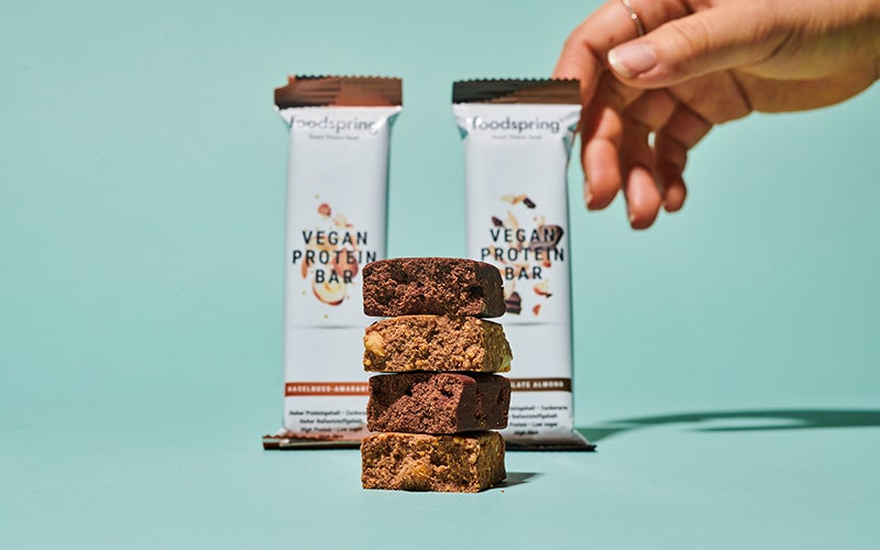 A dark-skinned hand reaches for a package of Vegan Protein Bar. There are 4 stacked pieces of the bars in the foreground of the picture.