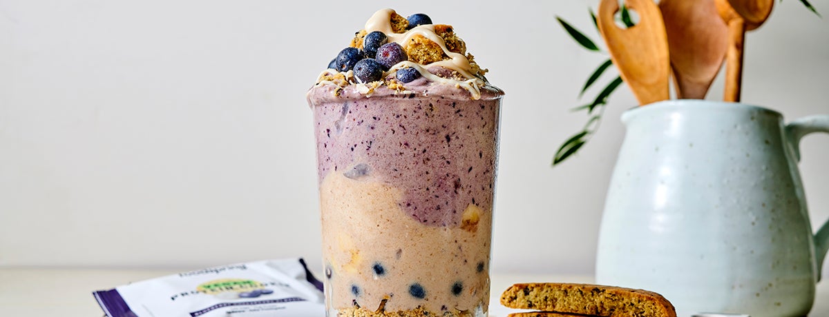 A big glass filled with blueberry cheesecake nice cream, topped with fresh blueberries and cookie crumbles