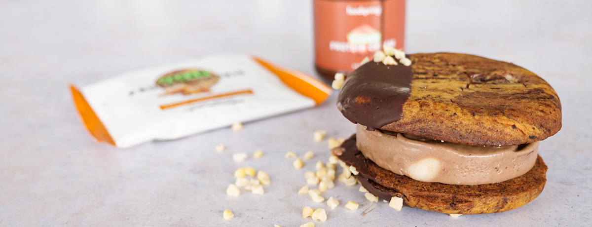 A vegan ice cream sandwich with dark chocolate over one side. The ice cream filling is a gentle beige. The cookies are a rich tan. There is a package of foodspring's Vegan Protein Cookies behind it.