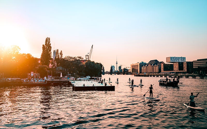 A riverscape at sunset. The TV Tower of Berlin is visible in the background, while a group of eight to ten people on the water use their stand up paddle board.