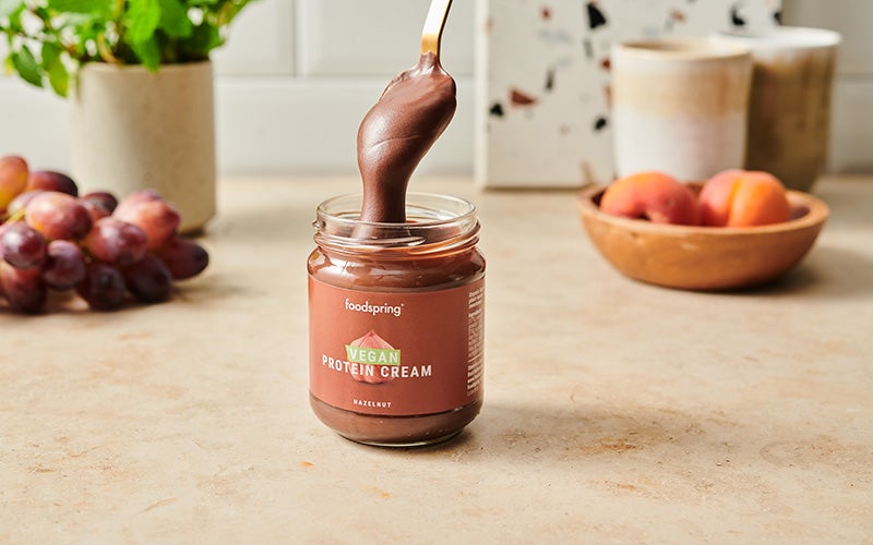 A golden spoon pulls a rich brown serving of vegan protein cream out of its glass jar