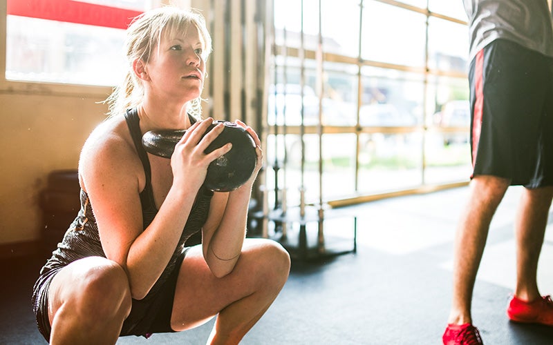 A blonde-haired, backlit, white woman holds a kettlebell while performing a goblet squat inside a gym