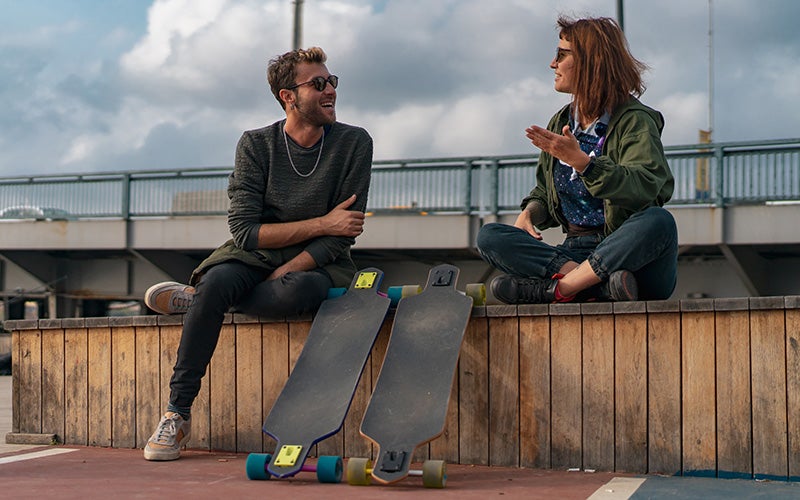 A white man and a white woman take a break from longboarding, resting their boards against a wooden divider wall which they are sitting on. They are chatting animatedly and smiling.