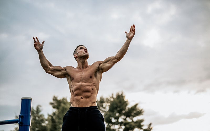 A shirtless white man with an upper body full of defined muscles raises his arms towards the sky. 