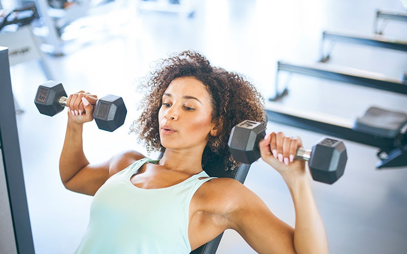 A woman of color performs exercises in her upper body workout for women. Here she is doing an incline press with dumbbells.