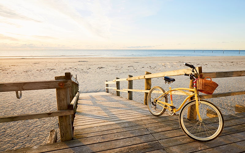 A yellow bike leans against the fence of a wooden boardwalk and ramp down to a light and sandy beach. The ocean beyond looks calm and still. The sky is light blue, and the sun is setting from off-camera, lending long shadows to the ground and a yellow-white glow to the left half of the sky.