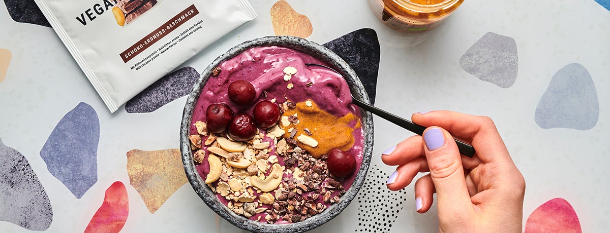 Are frozen foods healthy? Whip up this smoothie bowl and see how healthy they can be! In this photo, a white-skinned hand holds a spoon into a dark-cherry-pink smoothie bowl topped with cashews, cocoa nibs, a dollop of peanut butter, and fresh cherries.