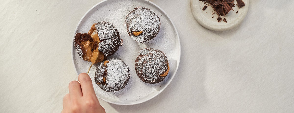 Salted caramel recipes for a salted caramel protein lava cake with a molten salted caramel core. A white hand holds a golden spoon, parting the powdered-sugar-dusted chocolate cake which enfolds a salted caramel center.