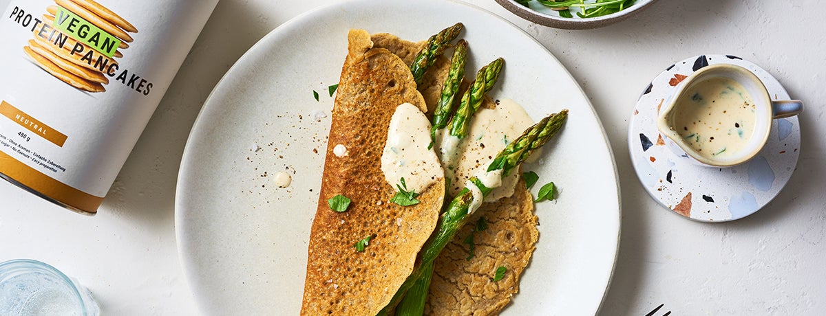 A white plate holds a folded-over vegan pancake with bright green asparagus. There is a small saucer of herb-specked hollandaise to the side.