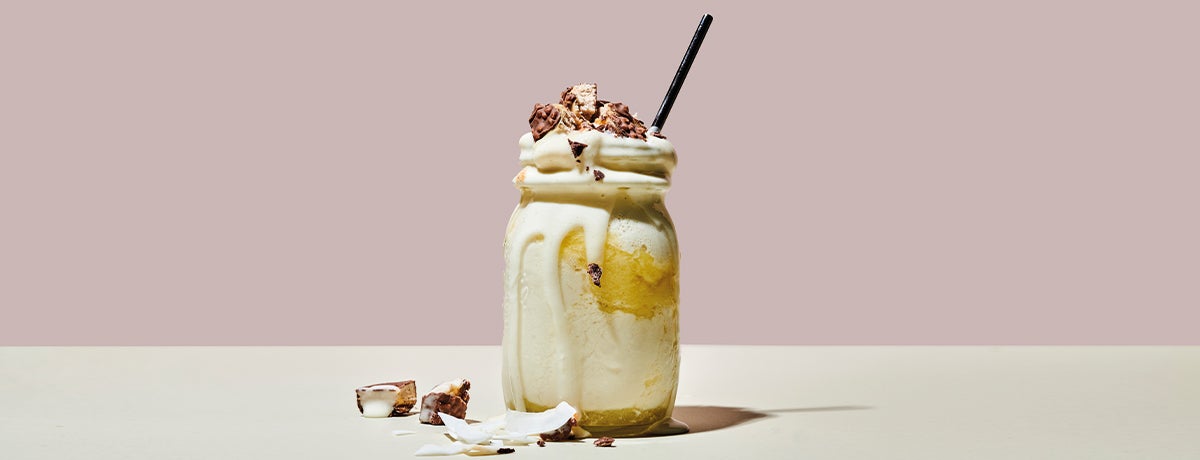 A Mason jar displays a summery yellow pina colada protein shake topped with crumbled Vegan Protein Bar pieces and coconut meat
