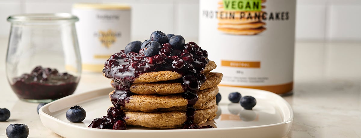 A stack of vegan blueberry pancakes topped with fresh blueberries and dripping over with blueberry jam