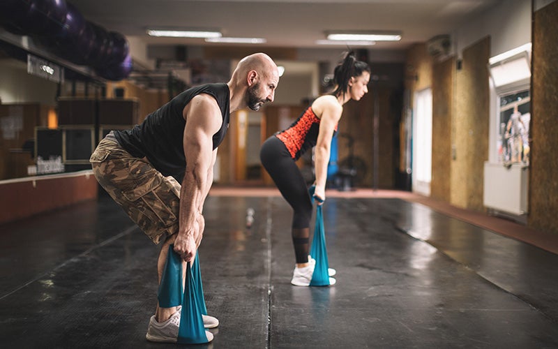 A white woman and white man do deadlifts with resistance bands indoors.