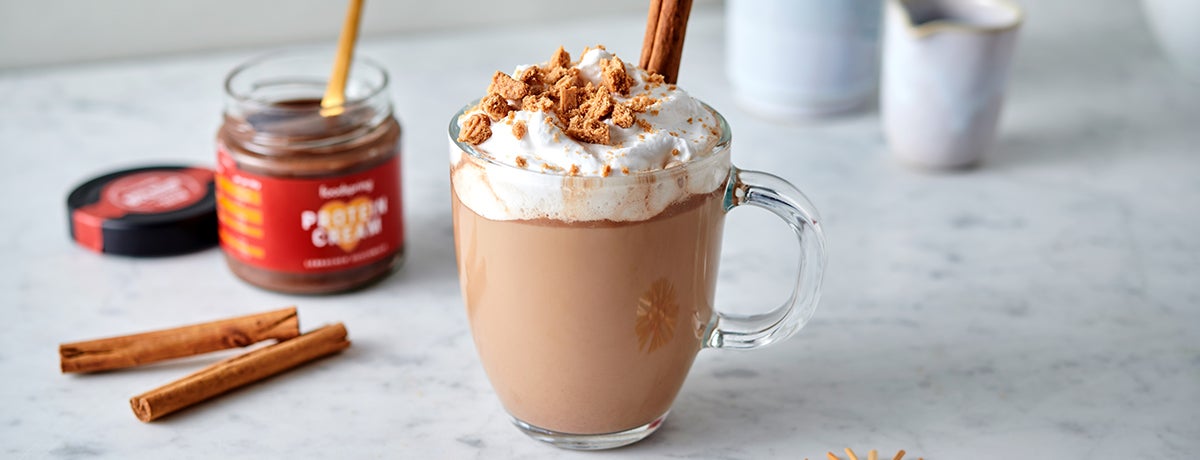 A glass of gingerbread latte topped with foamy whipped cream, with a jar of Gingerbread Protein Cream, cinnamon sticks, and other hot drinks in the background of the photo