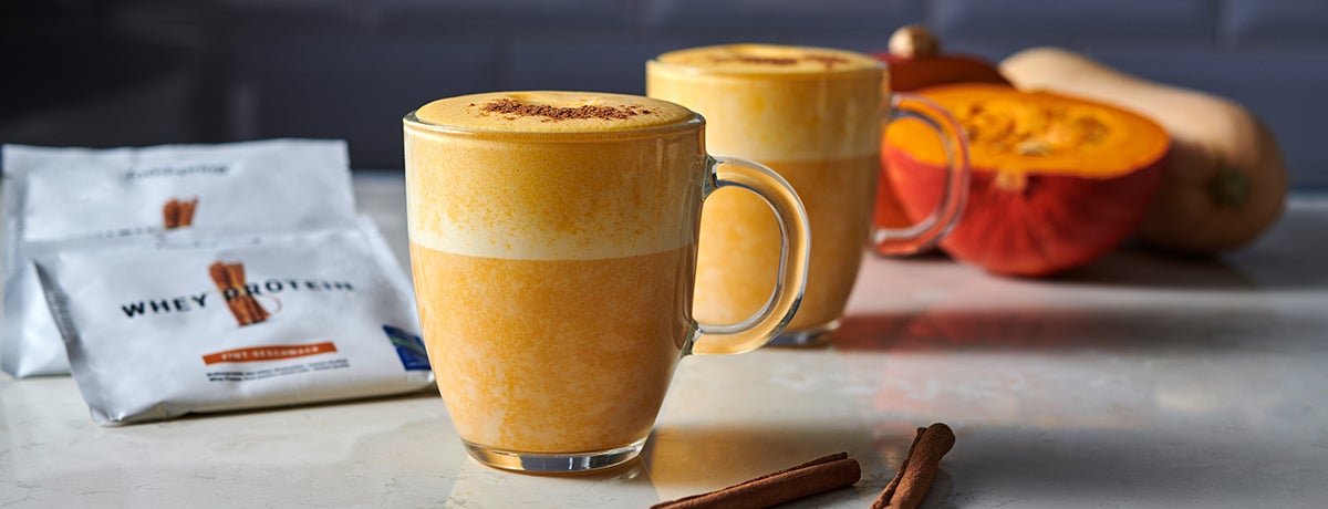 a glass holds a harvest-orange pumpkin spice latte with a packet of Whey Protein behind it