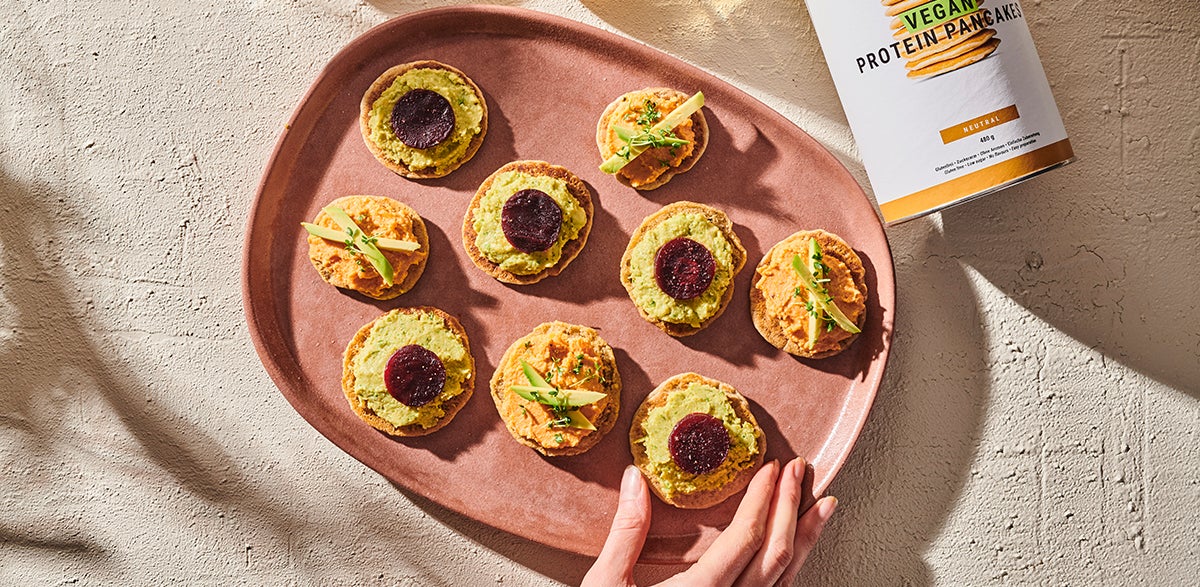 Vegan Blinis toped with either avocado wedges or purple carrot slices. Each blini is bite-sized and they rest on a terra-cotta plate as a white person's delicate-fingered hand reaches for one.
