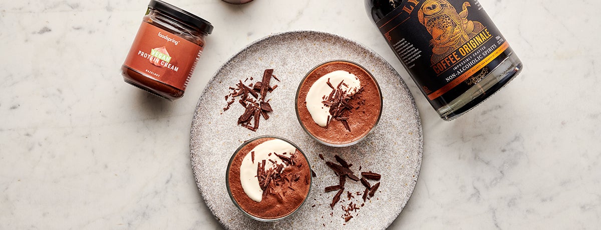Two bowls of vegan chocolate mousse sit on a plate. Each is topped with a dollop of cream and chocolate shavings. Next to the plate are displayed a jar of Vegan Protein Cream and a bottle of Lyre's Coffee Originale.