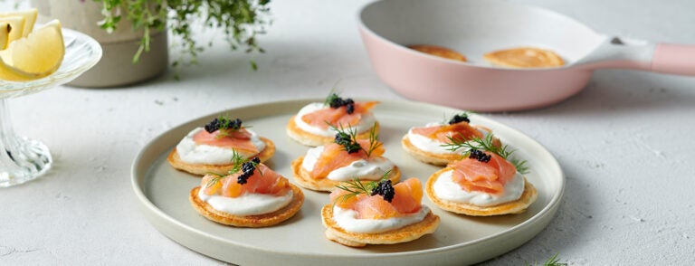 An off-white plate holds blinis topped with a dash of sour cream, a bite of smoked salmon, a pinch of caviar, and a spriglet of dill. One of many new year's recipes to impress your guests or yourself.