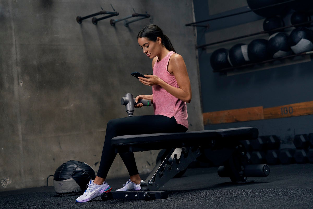 Women sitting on weight bench in gym using her phone and the Hypervolt 2