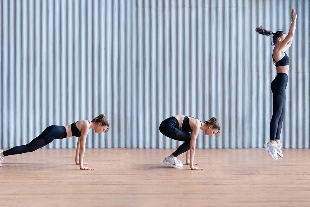 A thin white woman in sports bra and leggings does burpees to burn fat on a wooden floor with a corrugated light blue wall behind her