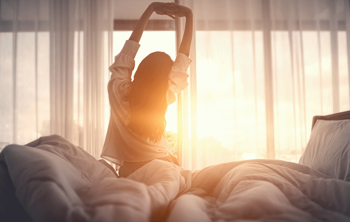 Frau streckt sich nach dem Aufstehen im Sonnenlicht vorm Fenster