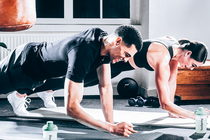 Man and women wearing black workout clothes doing planks with smiles on their face. Dumbbells and punching back in background.