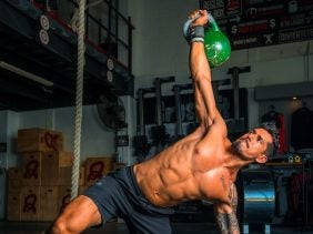 A man is working out with a kettle bell with six-pack abs. It looks like he is in the middle of a Turkish get up.