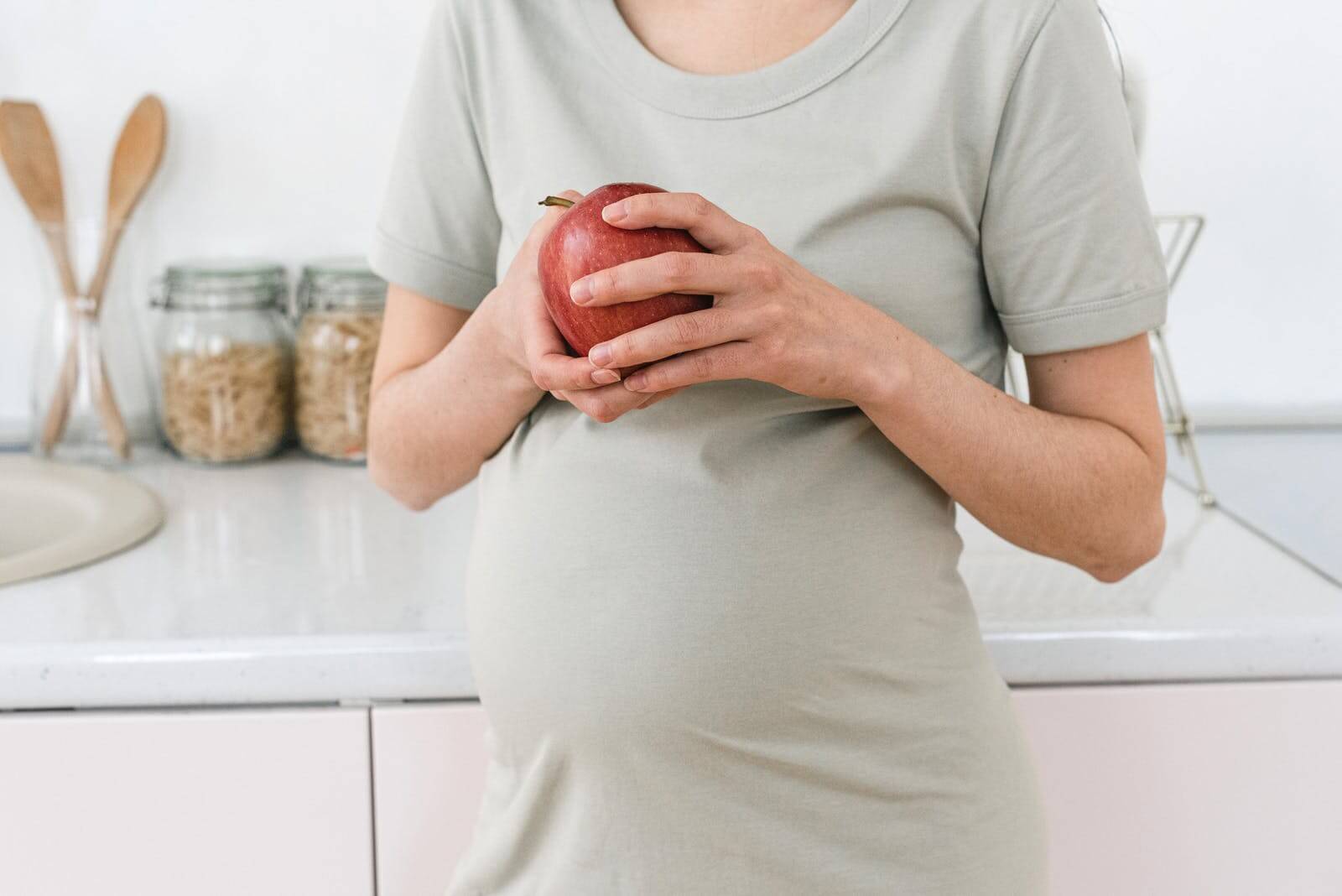 Eine schwangere Frau steht in einer Küche mit einem roten Apfel in beiden Händen direkt über ihrem Bauch