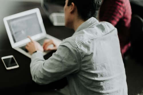 Ein Mann in hellem Hemd sitzt vor einem computer, links ein handy und rechts eine person in roter bluse