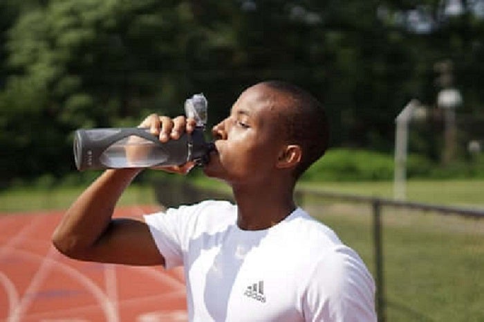 ein schwarzer Mann in einem weißen t-shirt trinkt Wasser aus einer Trinkflasche. Er steht auf eine Laufbahn mit grünem Hintergrund